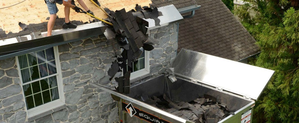 A person removing shingles from a house and loading the materials into a dumpster - How Long Does a Roof Tear Off Take?