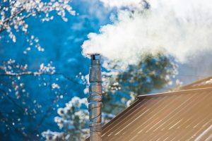 Smoke from the chimney on the roof during a sunny winter's day to illustrate Re-Roofing A Home In Winter Some Things For You to Consider