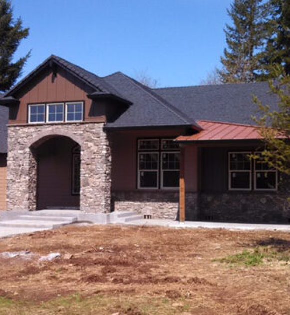 Custom home with dark architectural shingle roof, stone entryway, and covered front porch surrounded by trees.